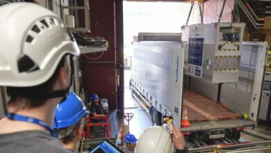 A view of a large white truck being loaded by crane with a large metal box. Workers wearing hard hats stand to the left of the truck.