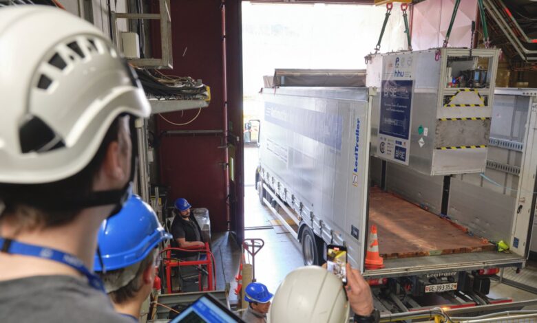 A view of a large white truck being loaded by crane with a large metal box. Workers wearing hard hats stand to the left of the truck.