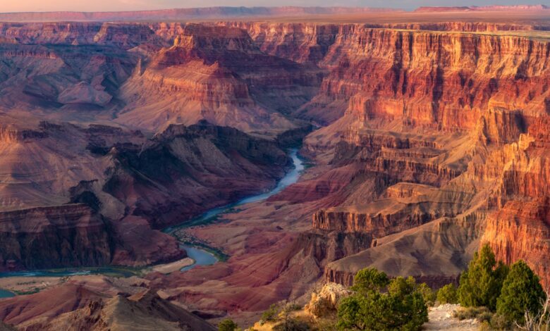 A series of flat red plateaus with a blue river cutting between them, creating a valley, with trees in the foreground.