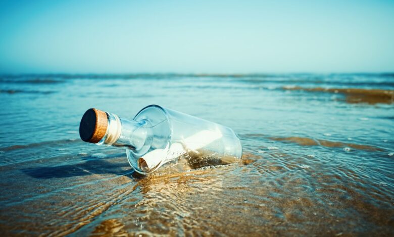 A large glass bottle with a cork holds a white rolled up piece of paper and sits on the beach.