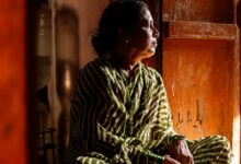 A woman in a green and white striped sari sits crosslegged in a wooden room, looking to the right of the camera