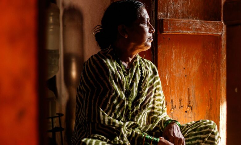 A woman in a green and white striped sari sits crosslegged in a wooden room, looking to the right of the camera