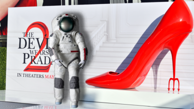 A larger poster for "The Devil Wears Prada 2" on a red carpet is in the background with an oversized red heel, in the foreground is a spacesuit.