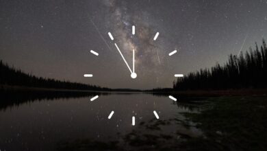 A stylized white clock face is superimposed over a night-time scene, in which meteors race through a starry sky over a lake bordered by the silhouettes of trees.