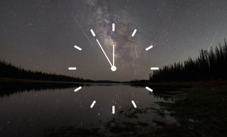 A stylized white clock face is superimposed over a night-time scene, in which meteors race through a starry sky over a lake bordered by the silhouettes of trees.