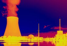 This infrared image shows a glowing orange nuclear power plant releasing hot yellow smoke near various outbuildings with blue water and sky above and below