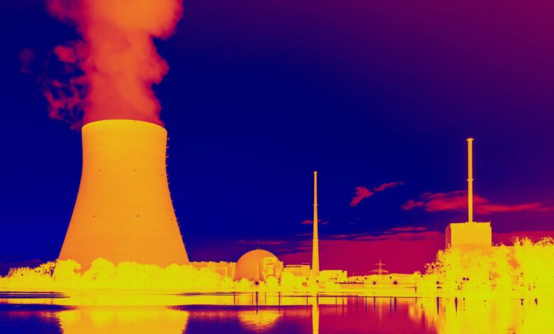 This infrared image shows a glowing orange nuclear power plant releasing hot yellow smoke near various outbuildings with blue water and sky above and below