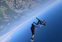 Two people hold hands while skydiving over Earth, a view of its surface next to the blue sky is seen.