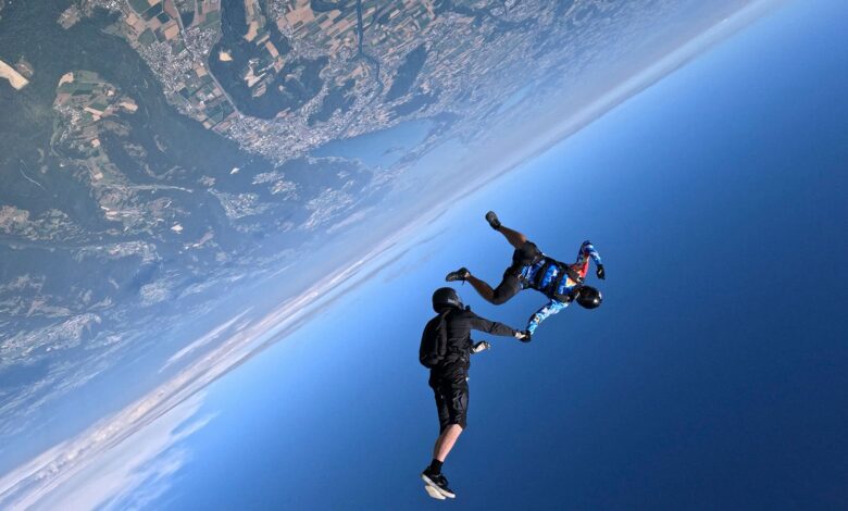 Two people hold hands while skydiving over Earth, a view of its surface next to the blue sky is seen.