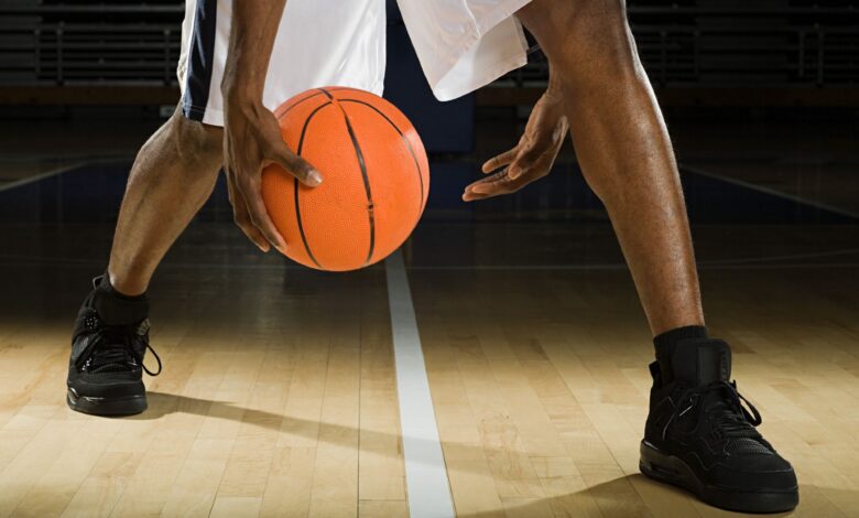 A close up of a man's lower legs, with both feet wearing tall black sneakers and black socks. He wears white basketball shorts and is bouncing a basketball between his hands while standing on a midline on a wooden basketball court